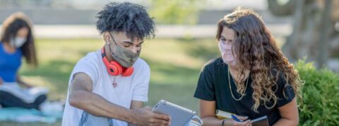 Two young poeple wearing face masks, young african american man on left showing young white woman an iPad, while outdoors