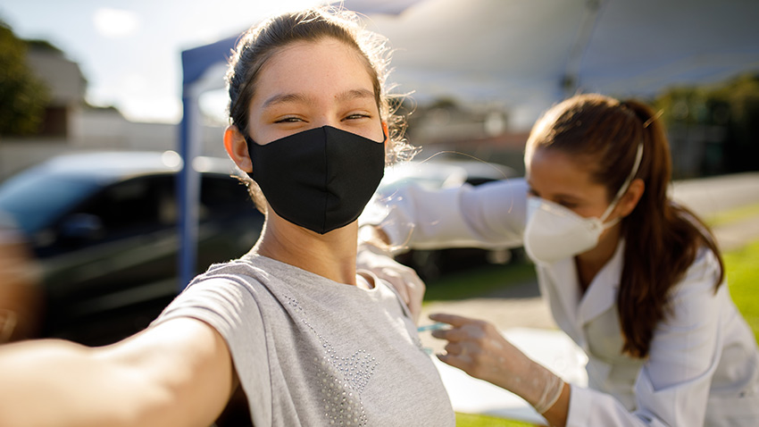 young woman smiling taking a selfie while wearing a face mask while a clinician administers a vaccine