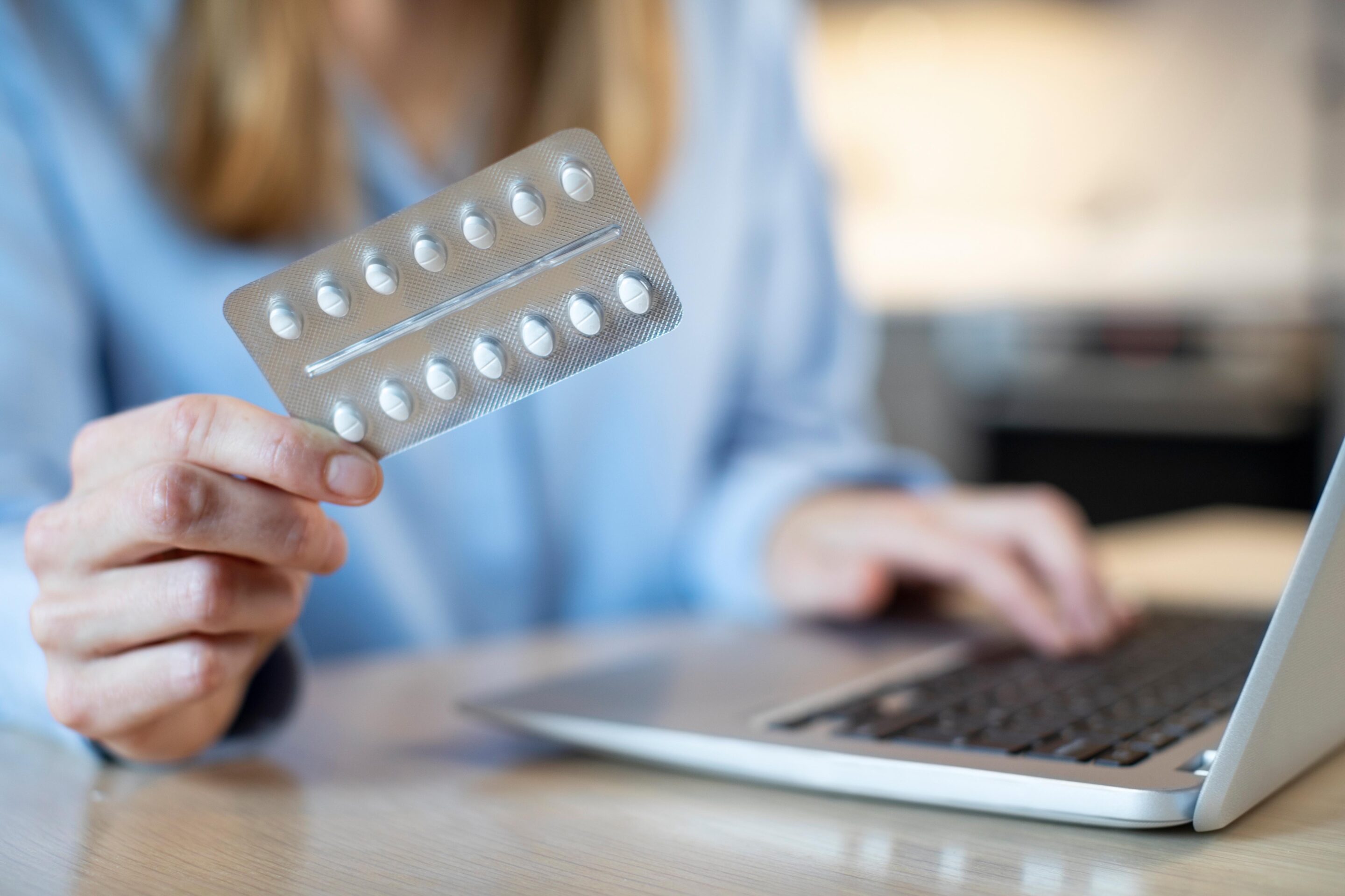 Close Up Of Woman At Home Looking Up Information About Medicatio Person holding a blister pack of white pills with a hand on a laptop computer keyboard.