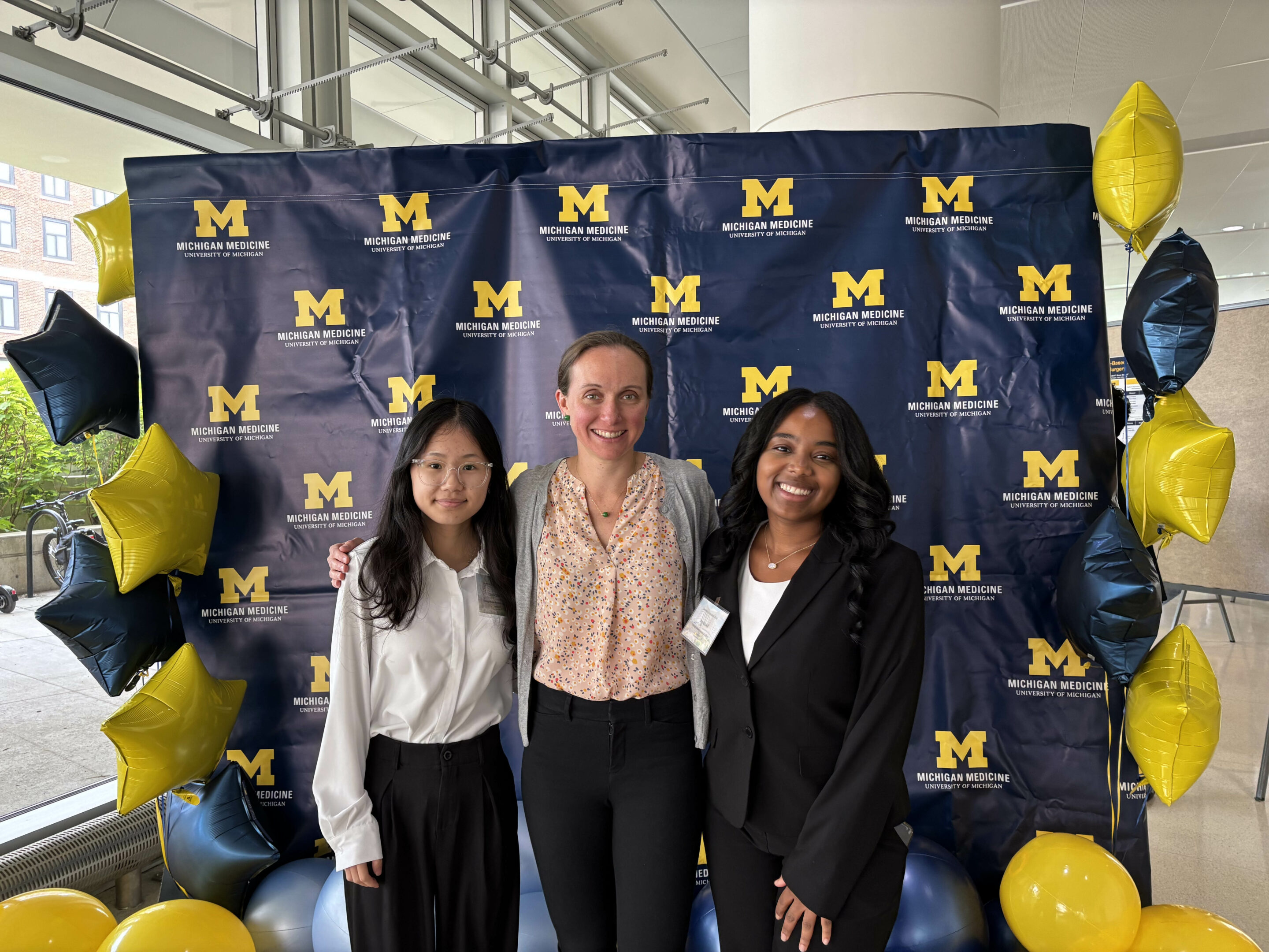 Group shot of three women of varying ages and races, standing in front of a screen that has the maize and blue University of Michigan logo on it. 