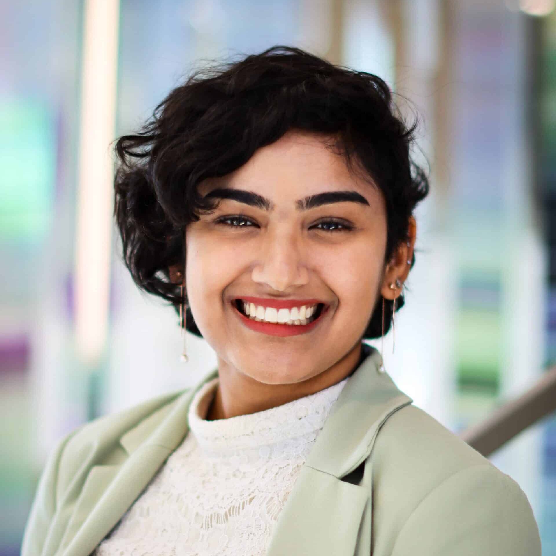 Young Indian woman with short, dark, curly hair, wearing a beige lapel collar coat and white lace top.