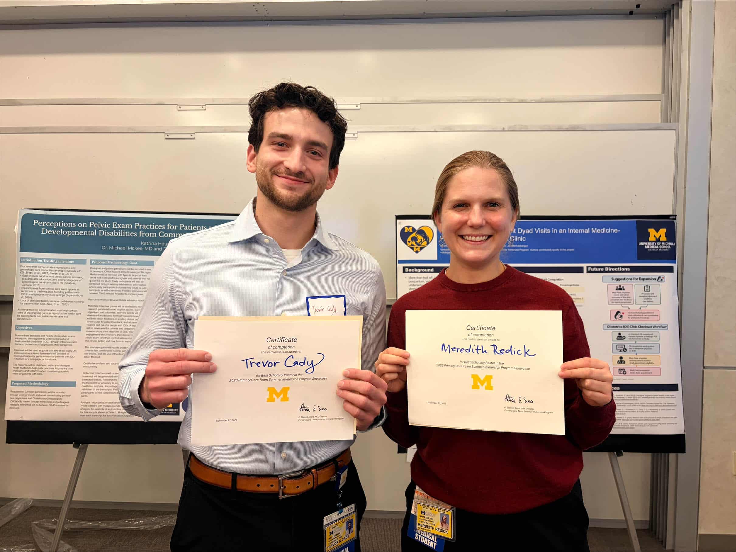 A young man with dark facial hair, wearing a button-down shirt, stands next to a young woman with dark blond hair, wearing a wine-colored sweater. They are both holding 'Best Poster' certificates and standing in front of two scientific posters on easels.